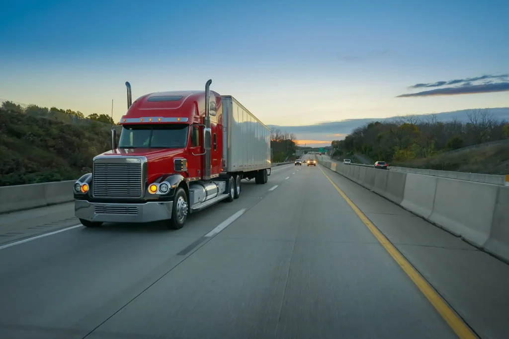 A red semi-truck drives along a divided highway at dusk, with a few cars visible ahead and trees lining both sides of the road under a blue and orange sky.