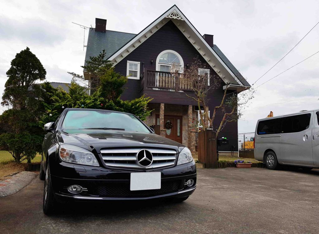 A black Mercedes-Benz car is parked in front of a dark-colored house with a gabled roof and balcony. A silver van is parked nearby, and trees and plants surround the house. The sky is overcast.