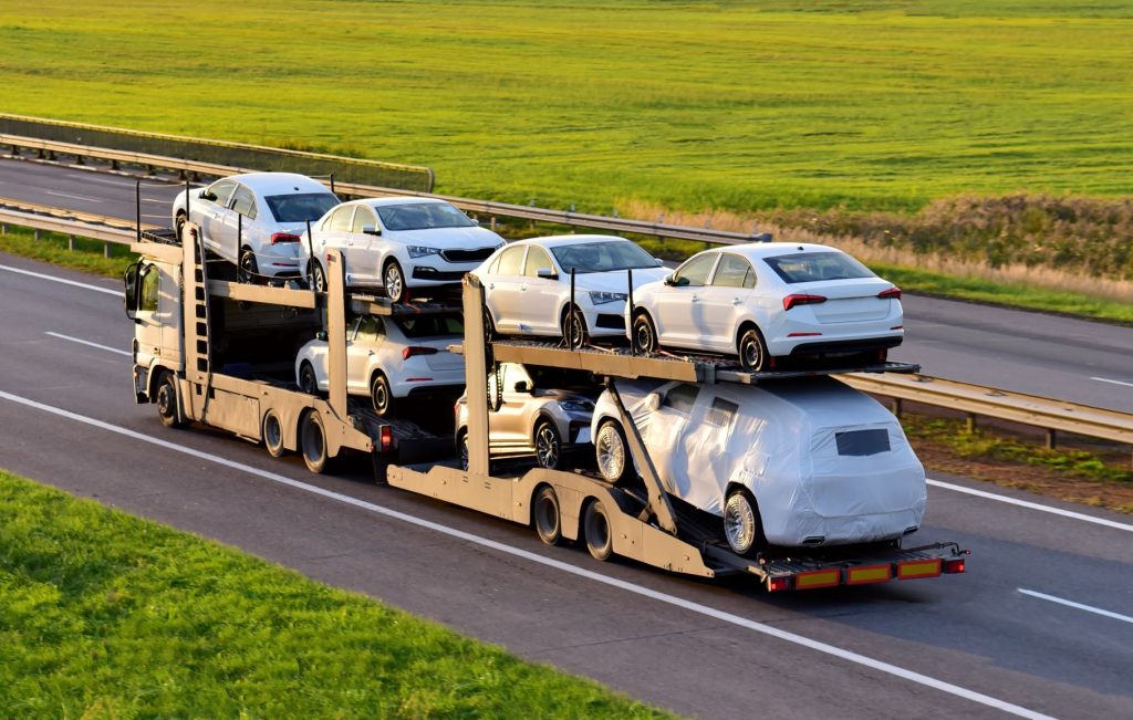 A car carrier truck transports several white sedans, with one car covered in a white protective wrap, on a multi-lane highway bordered by green grass and fields.